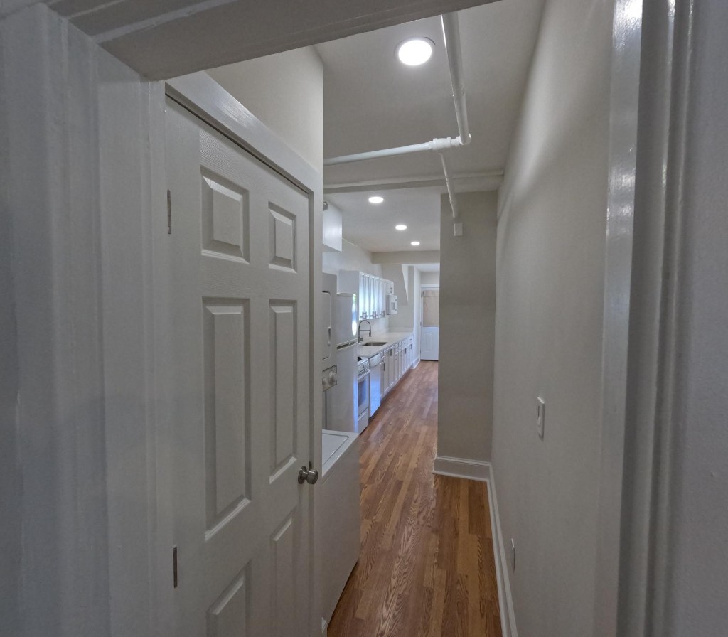 Hallway with hardwood floors leading to a galley kitchen, stacked laundry, and white cabinetry